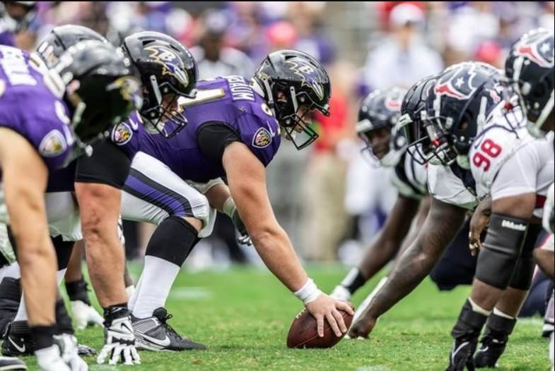 This image shows a football game between the Baltimore Ravens and the Houston Texans. The players are lined up at the line of scrimmage, with the Ravens in purple jerseys and the Texans in white jerseys